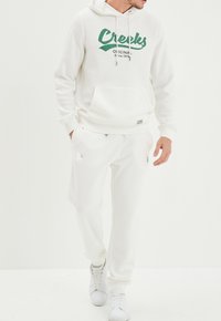 Witte hoodie met groen "Creeks" logo, kangoeroezak; gecombineerd met bijpassende witte sweatpants. Beide items zijn gemaakt van een zachte stof.