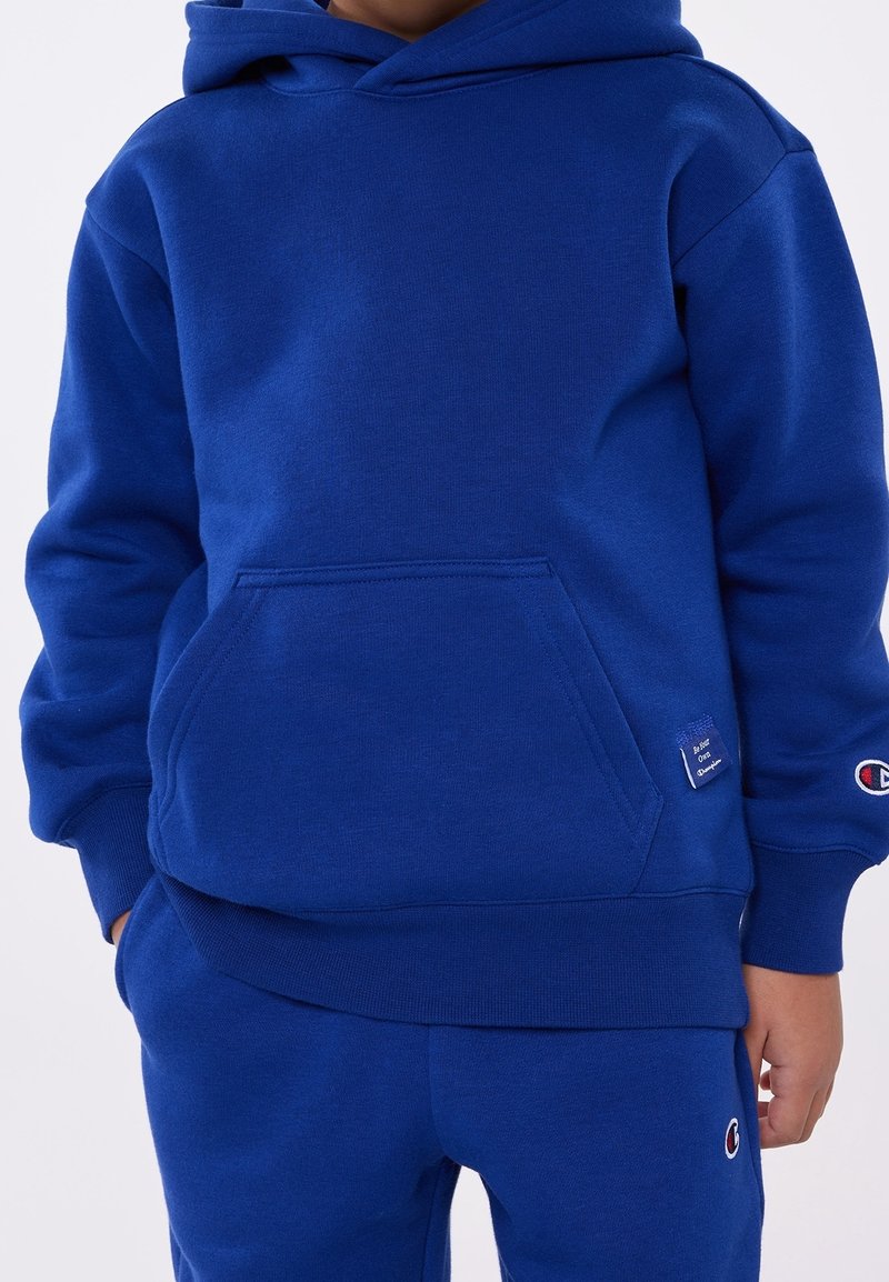 Enfant portant un sweat à capuche bleu avec poche avant et un pantalon de survêtement bleu assorti, tous deux arborant de petits logos Champion sur la manche, la poche et la jambe.