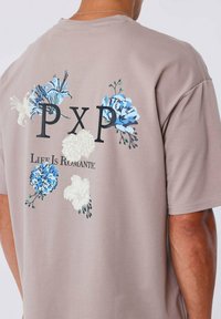 Camiseta de algodón gris con gráficos florales en azul y blanco, con el texto "PXP" y "La vida es romántica" impreso en la parte posterior. Mangas cortas.