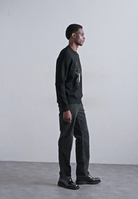Black cotton sweatshirt with green graphic detail, dark trousers, and black shoes. Side profile against a plain light backdrop.
