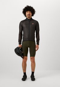 Lichte zwarte fietsjack met een hoge kraag en volledige rits. Olijfgroene shorts en zwarte sportschoenen. Man houdt een zwarte helm vast.