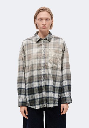 Jeune personne aux cheveux blonds courts portant une chemise à carreaux usée et trop grande boutonnée, ainsi qu'un pantalon sombre, debout sur un fond uni.