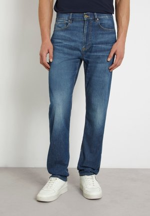 Jeans in denim blu con un design a gamba dritta, stile a cinque tasche e leggere scoloriture. Indossati con sneakers bianche su uno sfondo neutro.