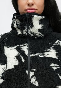 Μαύρο και άσπρο τζάκετ από φλίς με ψηλό γιακά, διακοσμημένο με υφή, κλείσιμο με φερμουάρ και μικρό λευκό λογότυπο Nike στο μπροστινό μέρος.