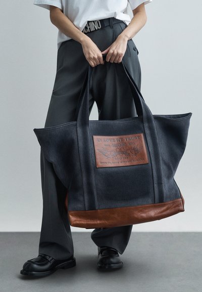 Άτομο που κρατάει μία μεγάλη καφέ και μαύρη τσάντα ώμου με δερμάτινη ετικέτα και κείμενο, φορώντας γκρι παντελόνι, λευκό πουκάμισο και μαύρα παπούτσια.