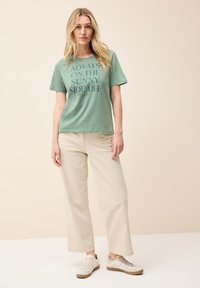 Camiseta verde menta con "Siempre del lado soleado de la vida" impreso en negrita, combinada con pantalones de pierna ancha beige y zapatillas de colores claros.