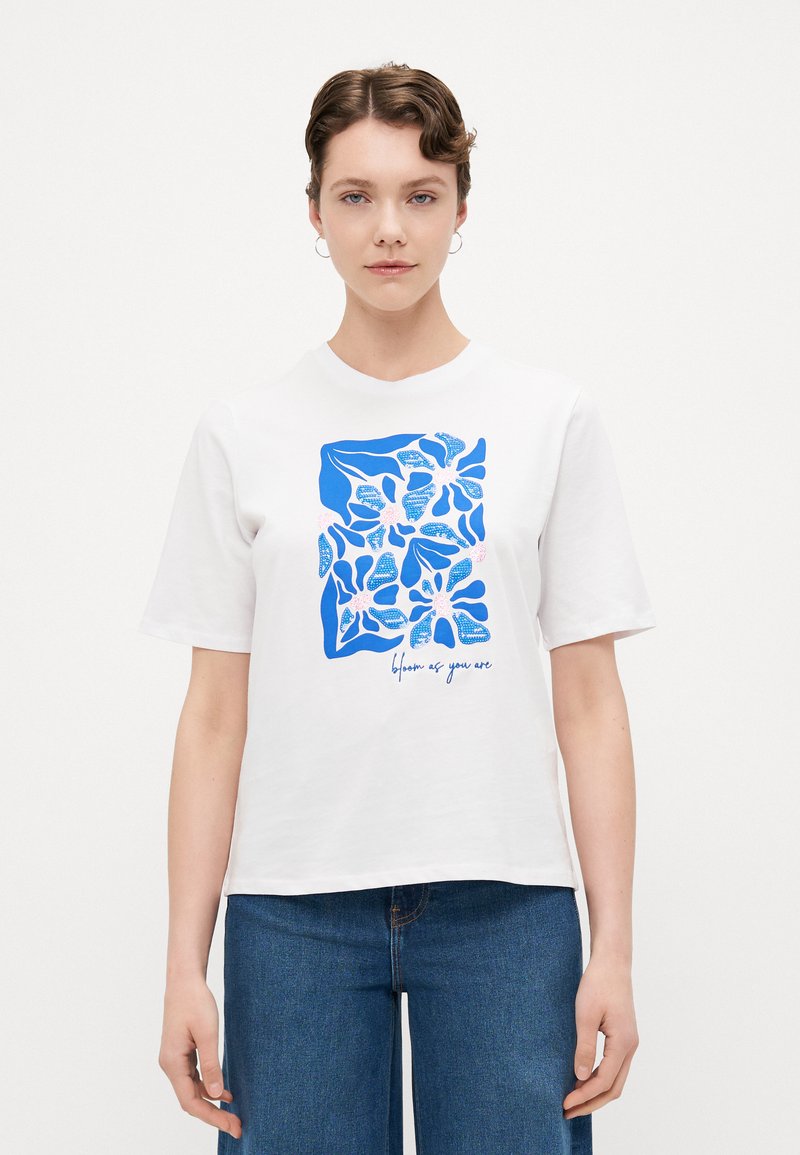 Witte katoenen t-shirt met een blauwe bloemenprint en de tekst "bloei zoals je bent." Korte mouwen en een ontspannen, boxy pasvorm.