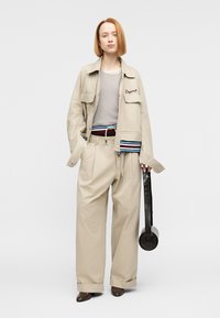 Μπεζ φαρδιά ζακέτα με κεντημένο κείμενο, γκρι ριμπ μπλουζάκι, φαρδιά παντελόνια με ζώνη, σκούρα καφέ παπούτσια και στρογγυλή μαύρη τσάντα.