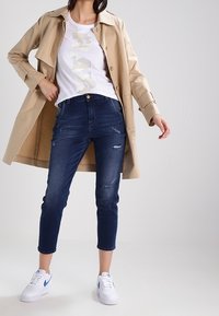 Beige trenčkot přes bílé tričko s camo potiskem, spárovaný s tmavě modrými zkrácenými džíny a bílými teniskami s modrými akcenty.