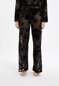 Zwarte doorschijnende broek met florale fluwelen patronen, gedragen met een bijpassende doorschijnende top, model staat blote voeten tegen een witte achtergrond.