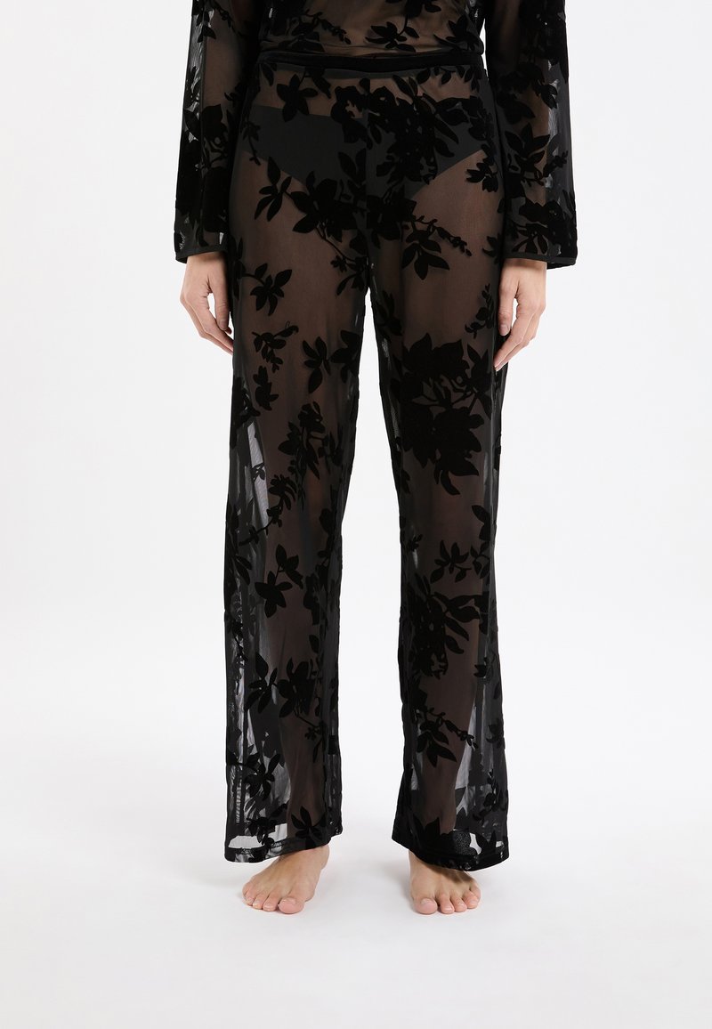 Zwarte doorschijnende broek met florale fluwelen patronen, gedragen met een bijpassende doorschijnende top, model staat blote voeten tegen een witte achtergrond.