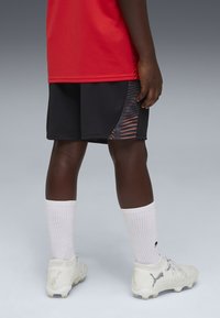 Sorte atletikshorts med orange mønsterdetaljer, kombineret med hvide sokker og hvide fodboldstøvler. Rød skjorte synlig bagfra.