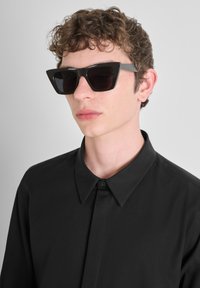 Gafas de sol geométricas negras con montura angular y lentes oscuros, llevadas con una camisa negra de botones, que presenta un collar en punta.
