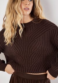 Brauner, gestrickter, verkürzter Pullover mit einem strukturierten Wellenmuster sowie geripptem Halsausschnitt und Bündchen. Kombiniert mit einem passenden braunen Rock.