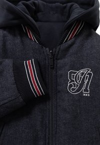 Donkergrijze herringbone patroon jas met capuchon, zwarte rits, ribgebreide manchetten met rode en witte strepen, en een wit geborduurd logo op de borst.