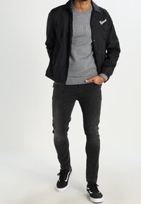 Chaqueta negra ligera con botones de presión, sudadera gris con texto, jeans negros ajustados y zapatillas negras con acentos blancos.