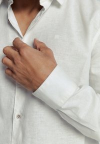 Witte linnen blouse met een klassieke kraag, voorzien van een knoopmanchet en een textuurstof. Een hand die de kraag aanpast is zichtbaar.