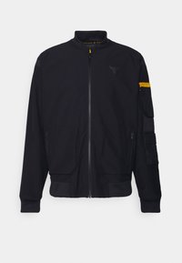 Svart bomberjacka i slät tyg, med dragkedja fram, ribbade manschetter, två framfickor och en ficka med dragkedja på ärmen med gula detaljer.