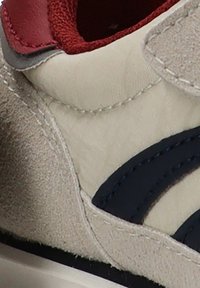 Zapatillas de ante beige con acentos curvados en azul oscuro, detalles en cuero rojo y suela de goma blanca. Presenta costuras cosidas y una superficie texturizada.