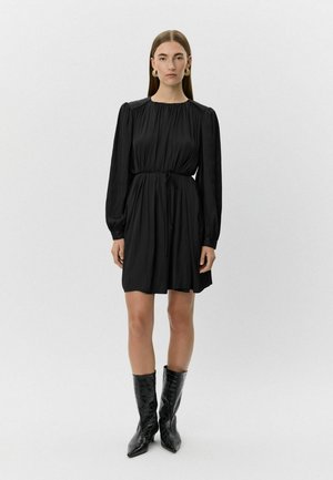 Juodas suknelė su susiraukšlėjusiu liemeniu, ilgomis rankovėmis ir minkšta, tekstūrine medžiaga. Sujungta su blizgiais juodais krokodilo rašto batais.