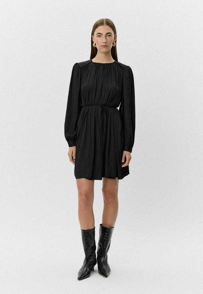 Juodas suknelė su susiraukšlėjusiu liemeniu, ilgomis rankovėmis ir minkšta, tekstūrine medžiaga. Sujungta su blizgiais juodais krokodilo rašto batais.