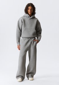 Personne aux cheveux bouclés portant un sweat à capuche gris ample et un pantalon de survêtement large assorti, debout avec une main dans la poche devant un fond uni.