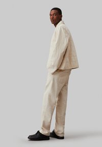 Giacca trapuntata color beige chiaro con colletto, abbinata a pantaloni beige coordinati. Calzature nere completano l'outfit, ambientato su uno sfondo grigio.