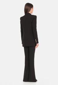 Blazer nero su misura con spalle strutturate, design monopetto e dettagli con bottoni dorati. Abbinato a pantaloni a zampa coordinati.
