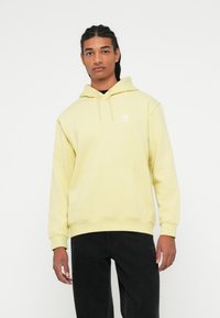 Mladenič z opletenimi lasmi, oblečen v svetlo rumen Adidas kapuco in črne hlače, stoji pred enobarvno belo ozadjem.