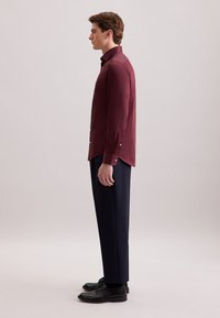 Bordeaux langemouwen shirt met een stevige textuur, iets getailleerde snit en witte knopen. Gecombineerd met donkere, op maat gemaakte broeken en zwarte schoenen.