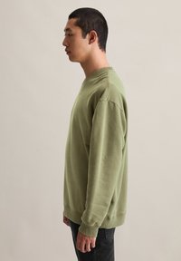 Jongere die zijwaarts staat in een olijfgroene sweatshirt en zwarte broek tegen een effen lichte achtergrond.