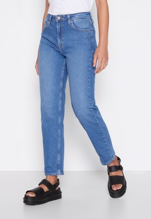 Personne portant un jean slim taille haute bleu et des sandales plateformes noires, debout sur un fond blanc.