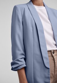 Blazer bleu clair avec des revers structurés, manches retroussées et tissu lisse. Porté sur une chemise blanche et un pantalon beige.
