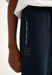 Marineblå sweatpants med en sort lynlås lomme, logodetalje og tekstureret stof. Elastisk talje med trykte grafikker på benet.