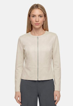 Femme aux cheveux blonds lisses portant une veste beige clair zippée et un pantalon gris foncé, debout devant un fond clair uni.