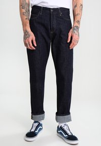 Dark denim jeans with a straight leg design, rolled cuffs, and yellow stitching. Paired with blue and white sneakers.