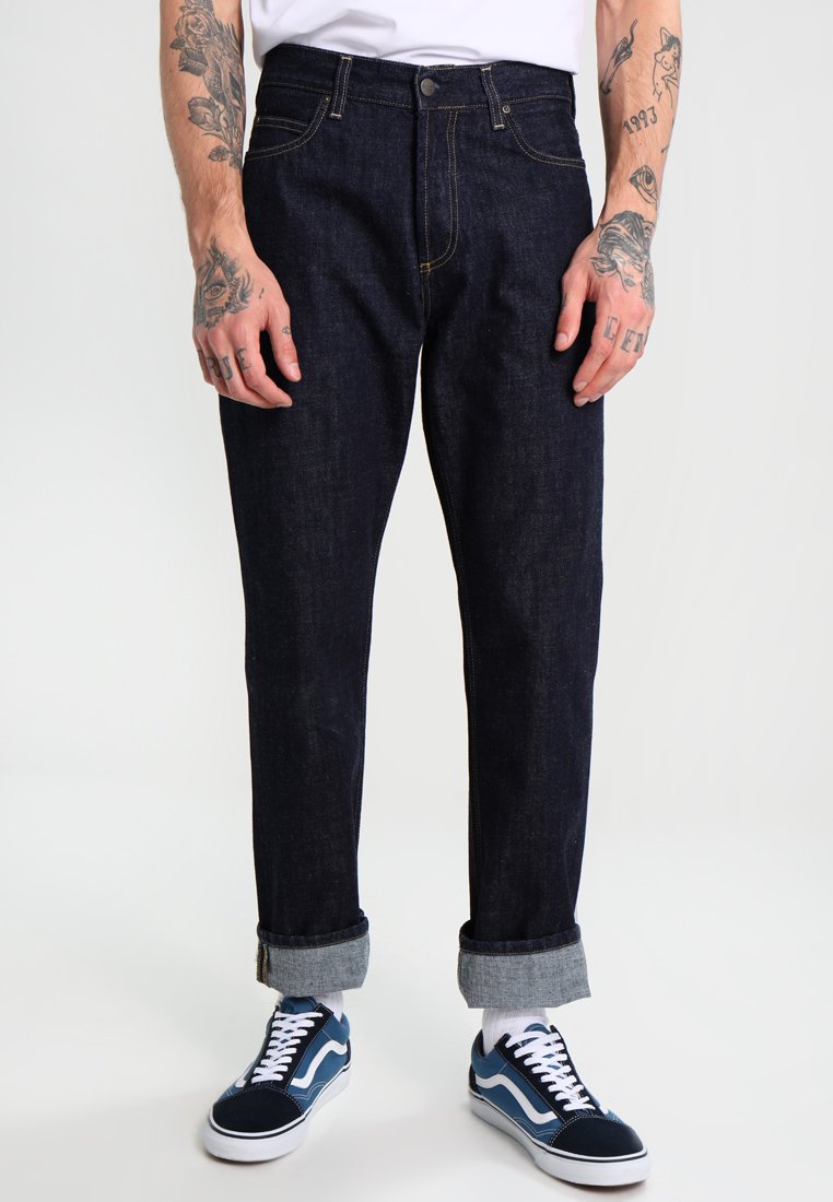 Dark denim jeans with a straight leg design, rolled cuffs, and yellow stitching. Paired with blue and white sneakers.
