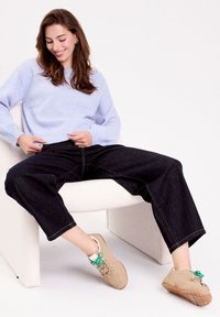 Un pull tricoté violet clair, un jean noir à jambes larges avec des surpiqûres jaunes, et des baskets beiges avec des lacets verts, assis sur une chaise blanche.