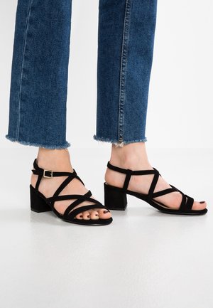 Schwarze Wildleder-Blockabsatzsandalen mit gekreuzten Riemen und einer Schnalle. Offene Zehenpartie mit glattem Finish und weicher Textur.