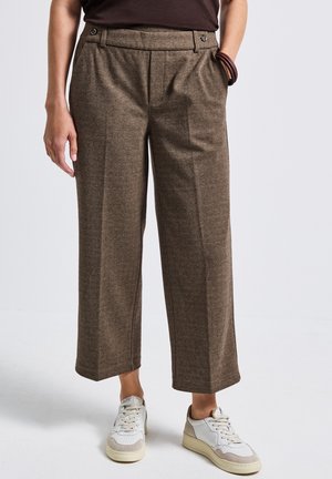 Kvinde iført brune brede cropped bukser, hvide sneakers og mørk top, med den ene hånd i lommen og armbånd om håndleddet.