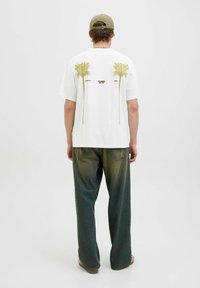Hvid kortærmet t-shirt med to grønne palmegrafikker på ryggen, parret med mørkegrønne, løstsiddende bukser.