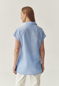 Světle modrá košile s krátkým rukávem a límečkem, s uvolněným střihem a rovním lemem. Měkká textilie, minimalistický design.