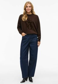 Sweatshirt marron avec texte graphique noir, associé à un jean large taille haute bleu foncé et des bottines noires. Tissu lisse et coupe décontractée.