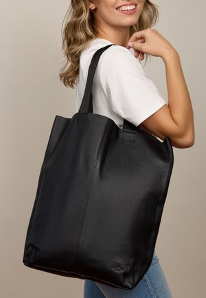Schwarze Ledertasche mit strukturierter Oberfläche, ausgestattet mit zwei stabilen Griffen und einem geräumigen Design, getragen von einer Person in lässiger Kleidung.