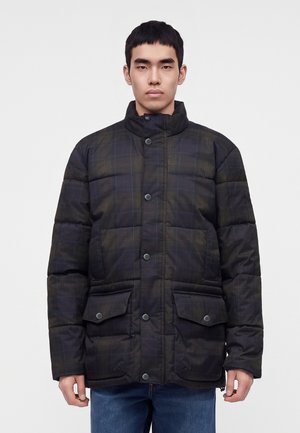Junger Mann trägt eine dunkle karierte gesteppte Jacke mit Druckknöpfen und vorderen Klappentaschen, kombiniert mit blauen Jeans, vor schlichtem Hintergrund.