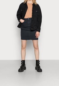 Chaqueta de mezclilla negra sobre una camiseta beige, combinada con una falda de mezclilla negra y botas negras gruesas con hebillas. Textura suave en todo.