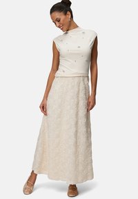 Robe couleur crème avec un haut fitted à manches courtes orné de décorations et une longue jupe texturée ornée de motifs floraux.