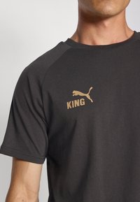Tamsiai pilkas trumpomis rankovėmis marškinėliai su auksiniu "PUMA" logotipu ir "KING" tekstu krūtinėje. Lygus audinys ir apvalus kaklas.
