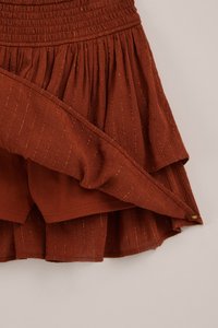 Ruosteenruskea kerroksellinen hame, jossa on kultaiset pystysuorat raidat ja joustava vyötärönauha. Sisemmässä pehmeässä kangasvuorissa ja ommelluissa helmassa yksityiskohdat näkyvissä.