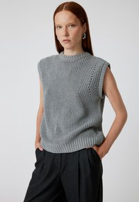 Maglione grigio lavorato a maglia senza maniche, con scollatura tonda, motivo testurizzato e dettagli ad occhiello laterali. Abbinato a pantaloni neri a vita alta.